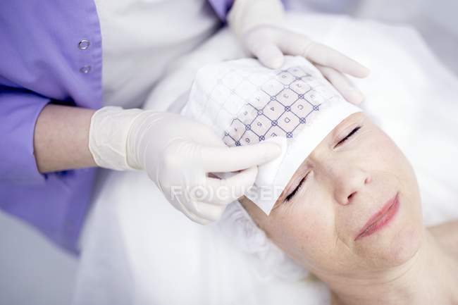 Dermatologist tracing grid paper on mature woman forehead for thermage therapy to soften wrinkles. — Stock Photo