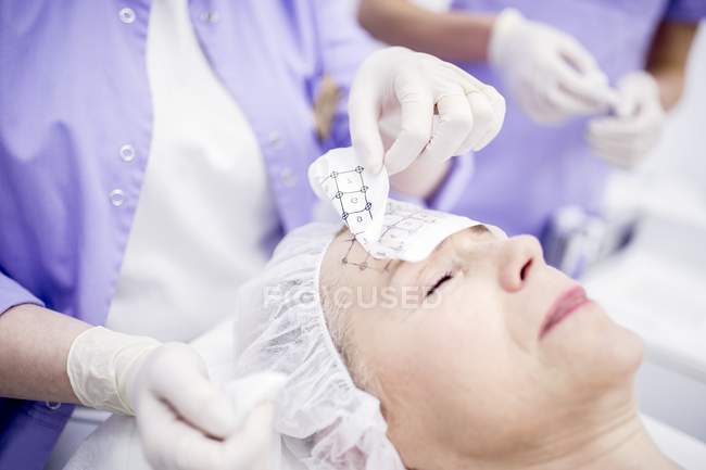 Dermatologist removing grid paper from mature woman forehead for thermage therapy to softening wrinkles. — Stock Photo