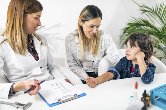 Female doctor performing allergy skin prick test on little boy with mother in immunologist office. — Stock Photo