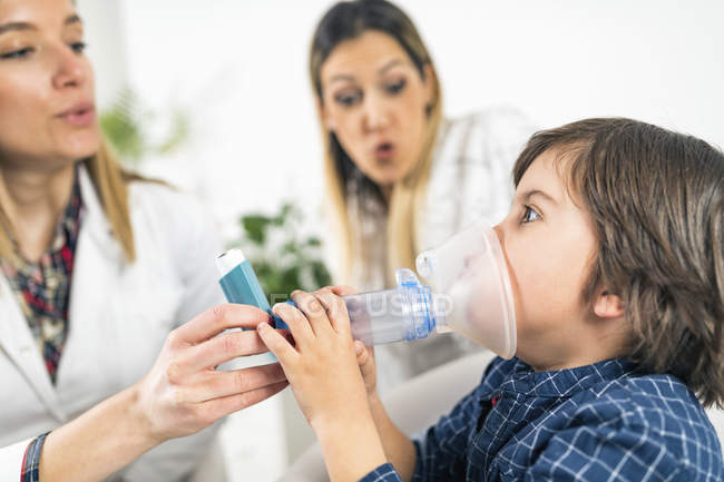 Pulmonologist helping little boy with inhaler, mother in background. — Stock Photo