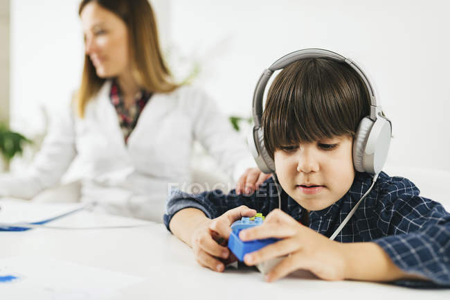 Niño usando auriculares como jugar con bloques de construcción durante la prueba de audición, médico femenino en segundo plano . - foto de stock