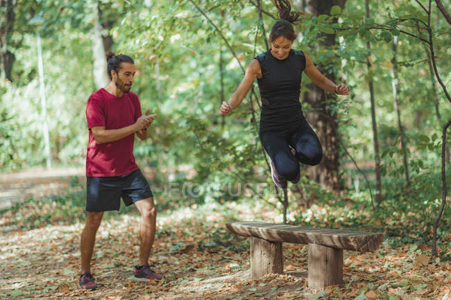 Young woman jumping on wooden bench with personal trainer while exercising outdoors. — Stock Photo
