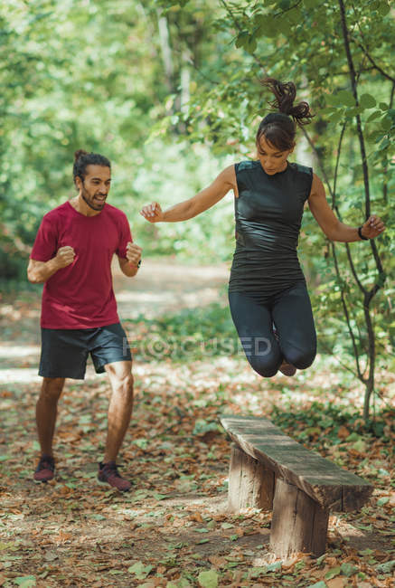 Young woman jumping over wooden bench while training with personal trainer in park. — Stock Photo