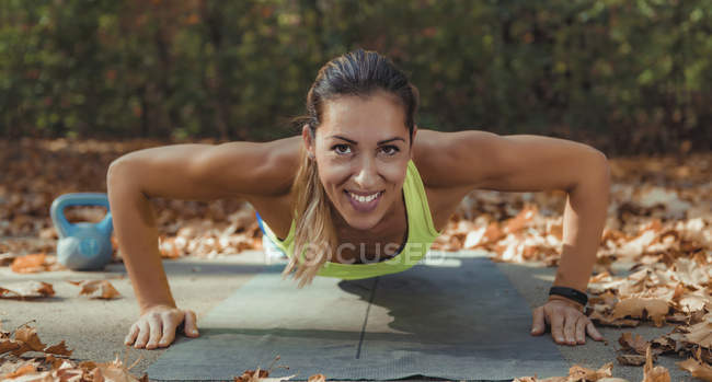 Lächelnde Frau, die in die Kamera schaut, während sie Liegestütze macht, während sie im Herbstpark im Freien Intensitätsintervall trainiert. — Stockfoto