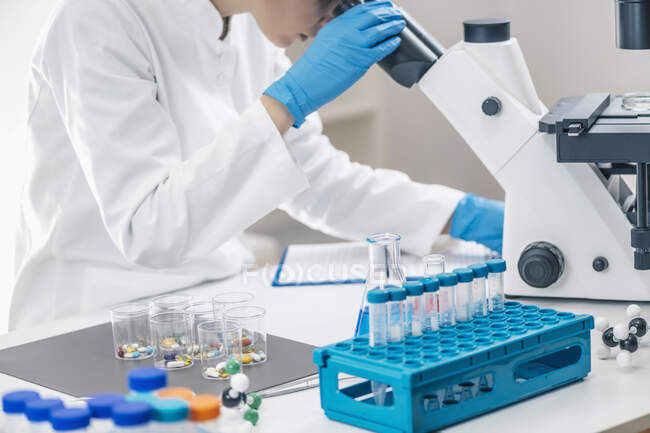 Investigador médico examinando un nuevo medicamento. estudiante de ciencias vestida con bata blanca de laboratorio mirando a través de un microscopio. - foto de stock