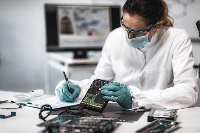 Female digital forensic expert examining computer hard drive and taking notes in police science laboratory. — Stock Photo