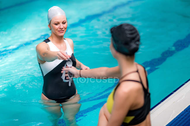 Giovani donne che consegnano occhiali a bordo piscina mentre riposano in piscina coperta . — Foto stock