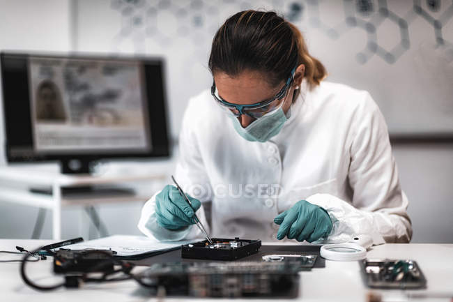Female police digital forensic science analyst examining computer hard drive with tweezers in lab. — Stock Photo