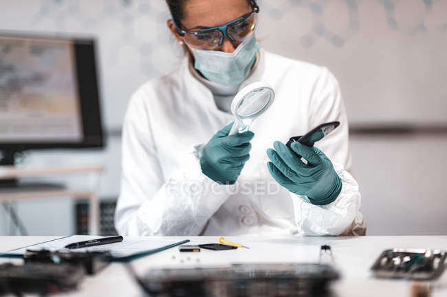 Police forensic analyst examining with magnifying glass confiscated mobile phone. — Stock Photo