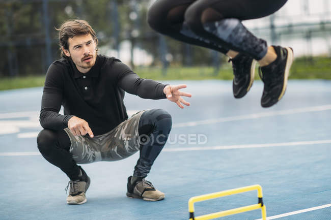 Sporty woman jumping over hurdle outdoors with fitness coach assistance. — Stock Photo
