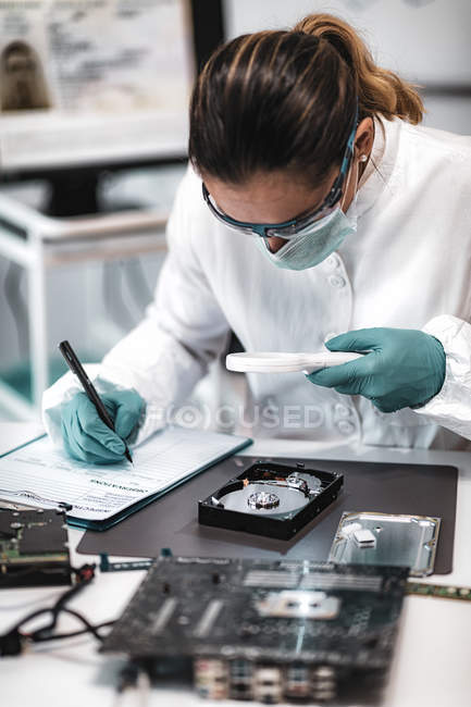 Tecnico forense digitale che esamina il disco rigido del computer con lente d'ingrandimento e prende appunti in laboratorio . — Foto stock