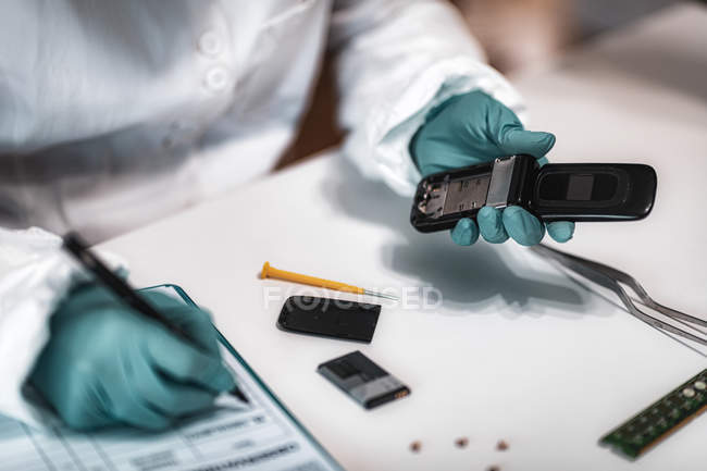 Эксперт-криминалист, изучающий конфискованный мобильный телефон и делающий записи в научной лаборатории
. — стоковое фото