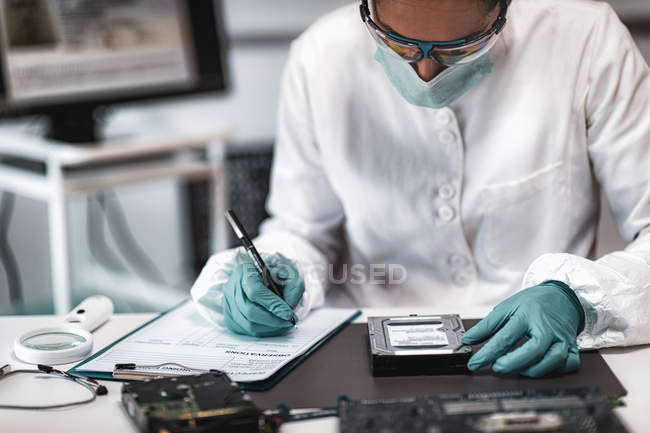 Female digital forensic expert examining computer hard drive and taking notes in police science laboratory. — Stock Photo