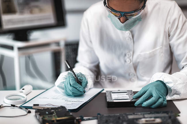 Experta forense digital femenina examinando el disco duro de la computadora y tomando notas en el laboratorio de ciencias de la policía . - foto de stock