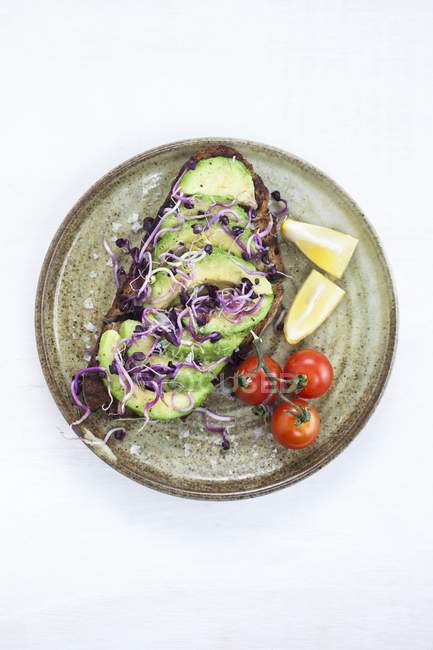 Lanche vegan saudável de abacate fresco em torradas com brotos, limão e tomate em prato redondo . — Fotografia de Stock