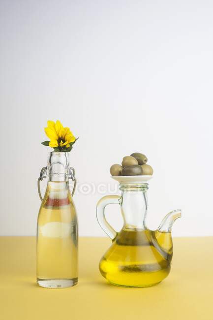 Bottle of sunflower oil and jug of olive oil with olives and flower, studio shot. — Stock Photo