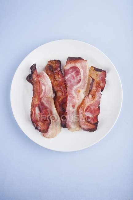 Bacon cozido na placa branca redonda no fundo azul . — Fotografia de Stock