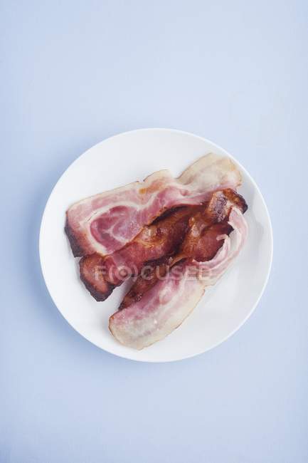 Bacon cozido na placa branca redonda no fundo azul . — Fotografia de Stock