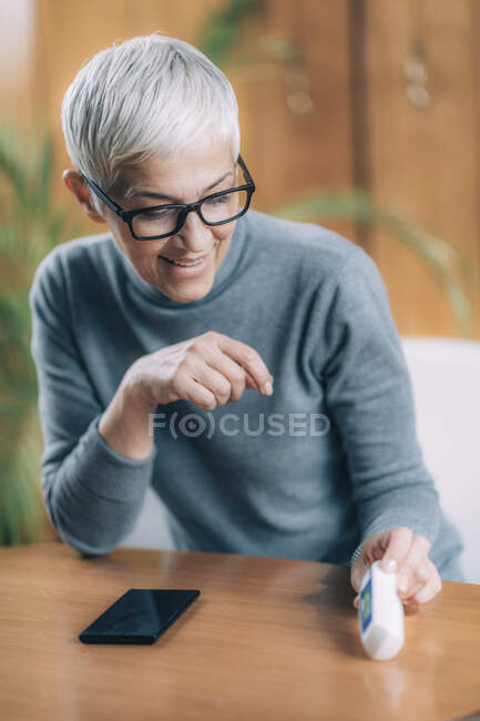 Mulher sênior gravando a temperatura corporal com termômetro digital sem contato, usando o aplicativo de telefone inteligente . — Fotografia de Stock