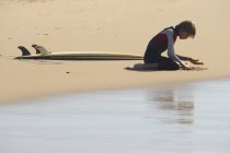 Garçon jouant au sable et assis à côté de la planche de surf — Photo de stock