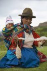 Cuzco, Perú, Mujer hilando lana de alpaca mientras lleva al bebé atrás - foto de stock
