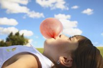 Fille couchée sur le dos et soufflant bubblegum en face du ciel bleu — Photo de stock