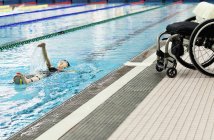 Паралитическая женщина плавает в бассейне на спине с инвалидной коляской на краю бассейна — стоковое фото