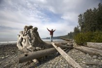 Hombre con los brazos levantados de pie en el tronco de deriva en Sombrio Beach, Vancouver Island, Columbia Británica, Canadá - foto de stock