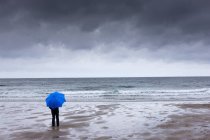 Uma pessoa segurando um guarda-chuva azul em pé na praia com um céu escuro sobrecarga; Northumberland, Inglaterra — Fotografia de Stock
