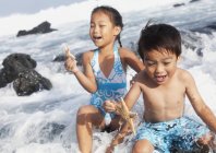 Un niño y una niña sostienen estrellas de mar y se sientan en las olas rompiendo en el borde del agua; Honolulu oahu hawaii estados unidos de América - foto de stock