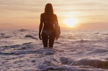 Жінка стоїть в океані і тримає дошку для серфінгу на заході сонця — стокове фото