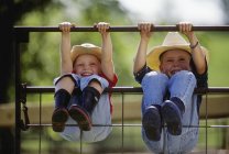 Young farm girl and boy wearing cowboy hats and rubber boots swing on metal gate, hanging by hands and legs — Stock Photo
