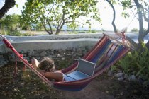 Fille à l'aide d'un ordinateur portable tout en étant assis dans un hamac — Photo de stock