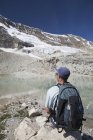 Randonneur assis sur un rocher avec un glacier sur le flanc d'une montagne coulant vers un étang de montagne réfléchissant avec ciel bleu ; Field, Colombie-Britannique, Canada — Photo de stock