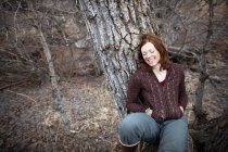 Retrato de una mujer con el pelo rojo apoyado en un árbol y sonriendo con los ojos cerrados - foto de stock