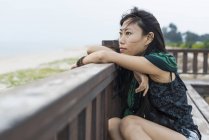 Jovem mulher sentada contra trilhos de madeira e olhando para o oceano — Fotografia de Stock