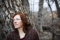Portrait d'une femme aux cheveux roux contre un arbre et regardant contemplatif — Photo de stock