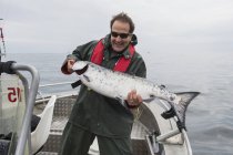 Человек стоит на рыбацкой лодке, держа недавно пойманного большого лосося. Острова Королевы Шарлотты, Британская Колумбия, Канада — стоковое фото