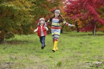 Мальчик и девочка в резиновых сапогах и разноцветной одежде бегут по полю, держа за руки деревья в ярких осенних цветах на заднем плане; Орегон, США — стоковое фото