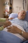Hombre mayor en cama de hospital cogido de la mano de la mujer - foto de stock
