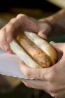 Salsicha de churrasco em um pão às mãos com fundo embaçado — Fotografia de Stock