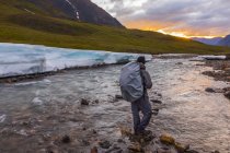 Uomo con zaino coperto che cammina sull'acqua di fiume di montagna con pietre contro colline sulla riva — Foto stock
