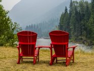 Dos sillas rojas de madera en la orilla sobre hierba contra el agua del lago con árboles y pendientes en la distancia - foto de stock