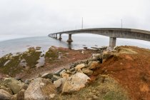 Plage de sable avec tas de pierres contre l'eau et pont sur l'eau pendant la journée — Photo de stock