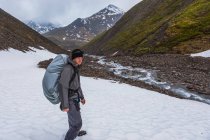 Mann mit gedecktem Rucksack steht über Bergfeld mit Schnee und Gipfeln im Hintergrund — Stockfoto