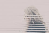 Femme aux cheveux blonds vu cacher son visage à travers une fenêtre avec des stores ; Connecticut, États-Unis d'Amérique — Photo de stock