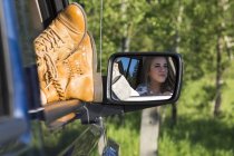 Vista delle gambe delle donne negli stivali sul bordo del finestrino delle auto e riflessione nello specchio contro gli alberi — Foto stock