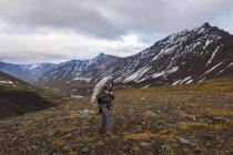 Homem com mochila coberta caminhando sobre o campo de montanha com plantas e picos no fundo — Fotografia de Stock