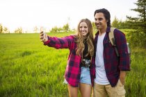 Pareja feliz de pie en el campo de hierba verde y hacer selfie en el teléfono inteligente - foto de stock