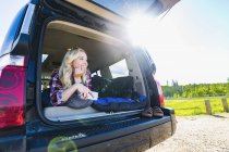 Chica joven rubia con gafas de sol en el coche y sonriendo mientras mira hacia otro lado - foto de stock